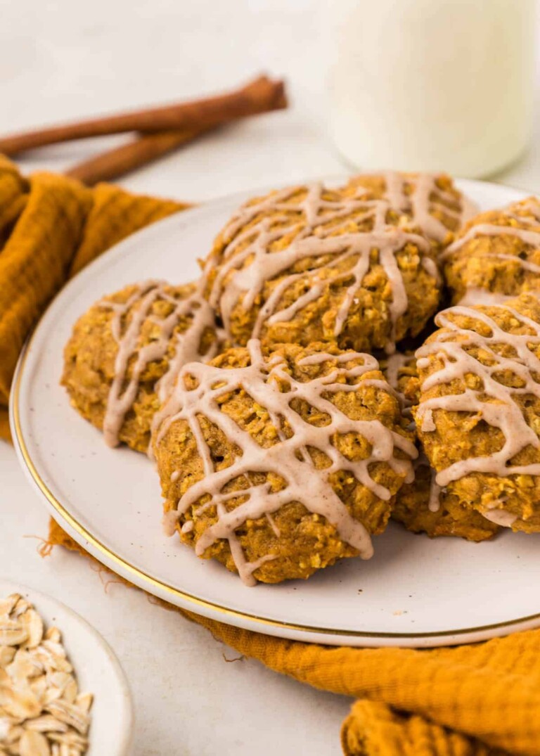 A plate of pumpkin oatmeal cookies drizzled with icing, set on a white surface with a yellow cloth, cinnamon sticks, and a small bowl of oats in the background.