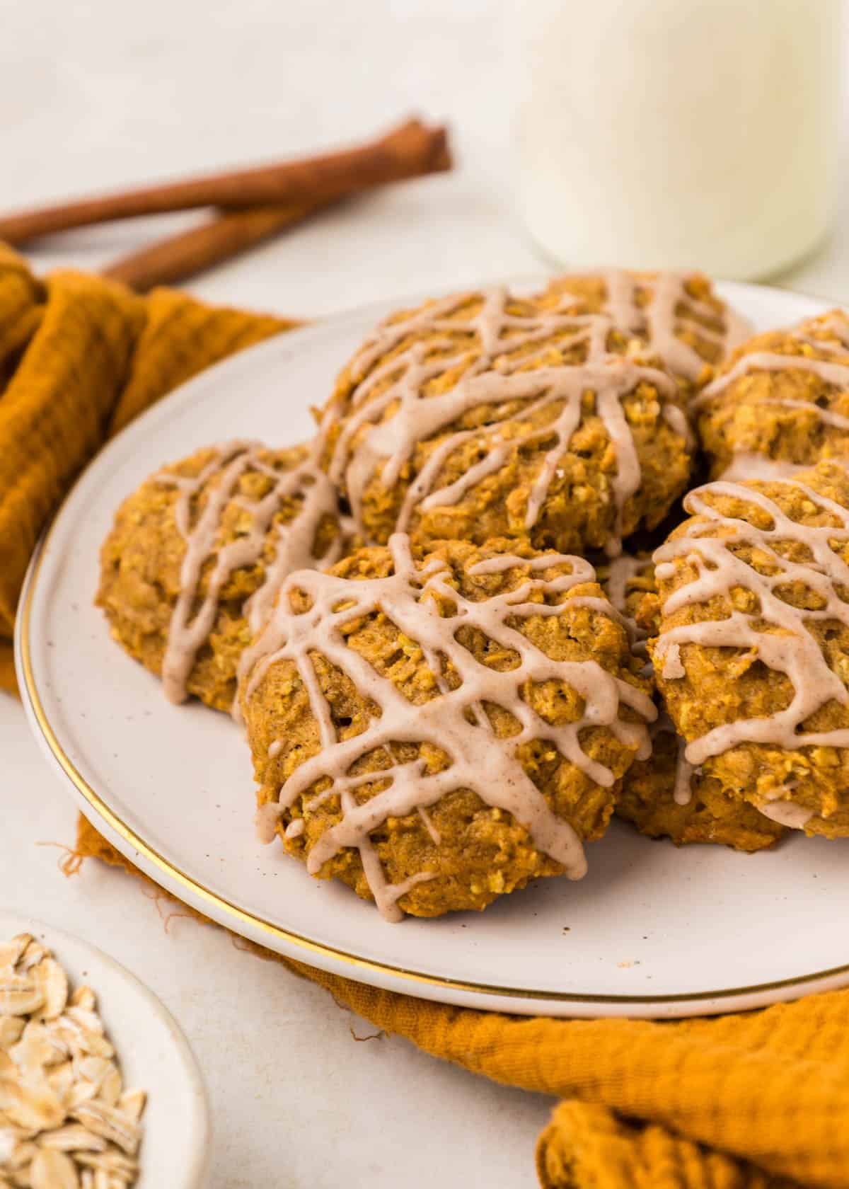 A plate of pumpkin oatmeal cookies drizzled with icing, set on a white surface with a yellow cloth, cinnamon sticks, and a small bowl of oats in the background.