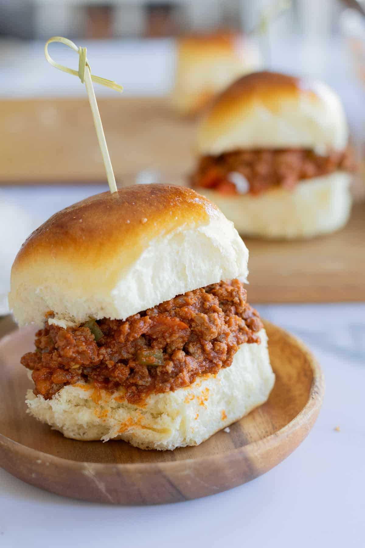 A close-up of a sloppy joe slider on a wooden plate, with another slider blurred in the background.