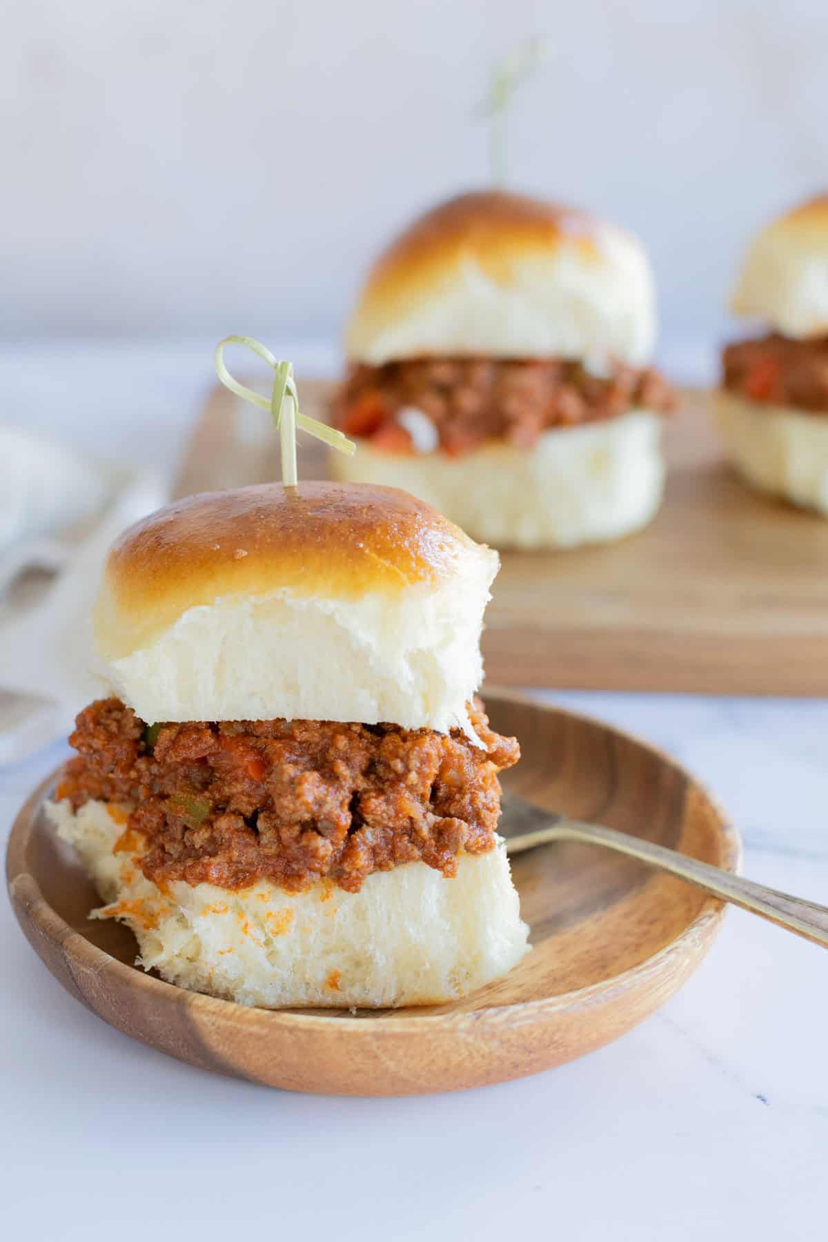 Three sloppy joe sliders are displayed, one on a small wooden plate with a fork, and two more in the background on a wooden board.