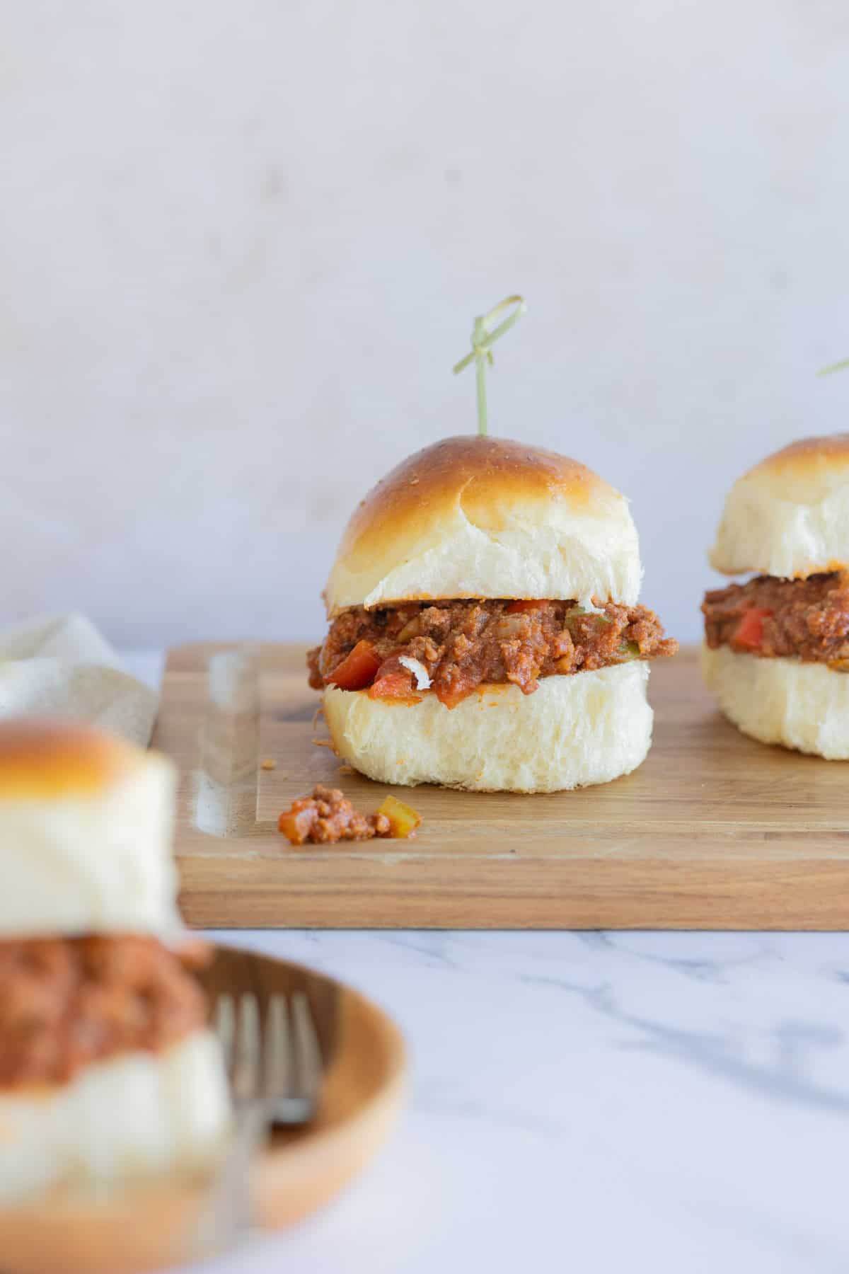 Two sloppy joe mini sliders placed on a wooden board, with a partial view of a third sandwich in the foreground. Each sandwich is topped with a round toothpick.
