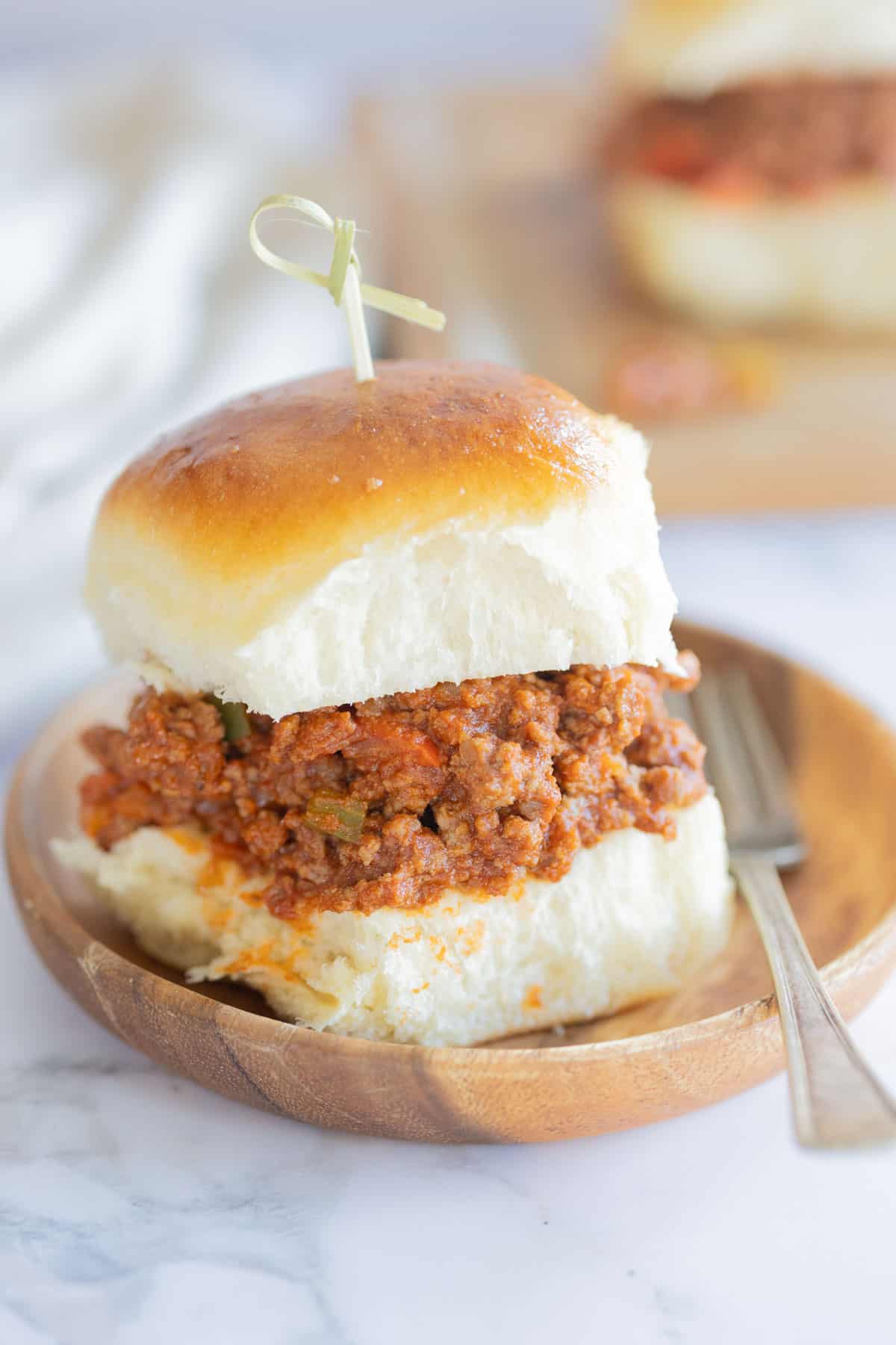 A sloppy joe slider topped with a small wooden pick, served on a round wooden plate with a fork beside it.