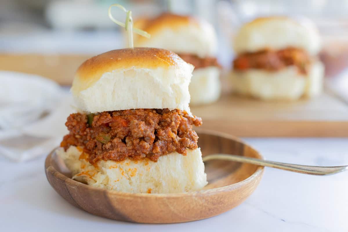 A sloppy joe slider on a wooden plate with a decorative skewer through the top bun. Two more sliders are blurred in the background on a wooden board.