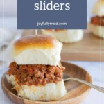 A sloppy joe slider on a wooden plate with a fork, featuring a soft bun filled with ground meat and sauce. Background includes another slider. Text above reads "Sloppy Joe Sliders" and "joyfullymad.com".