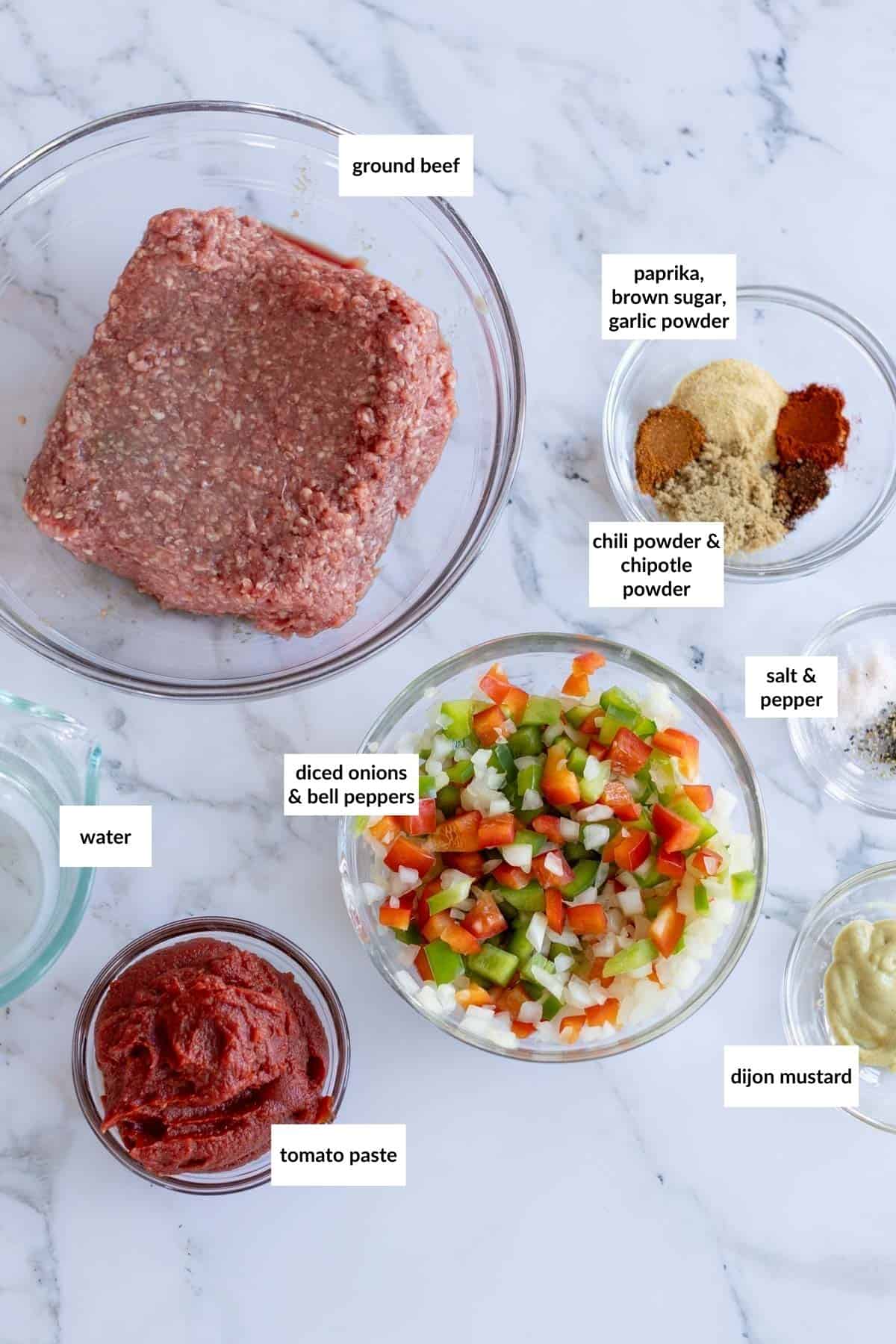 Various ingredients for cooking sloppy Joe sliders are laid out on a white marble surface, including ground beef, diced onions and bell peppers, tomato paste, mustard, water, and a mix of spices, each labeled.