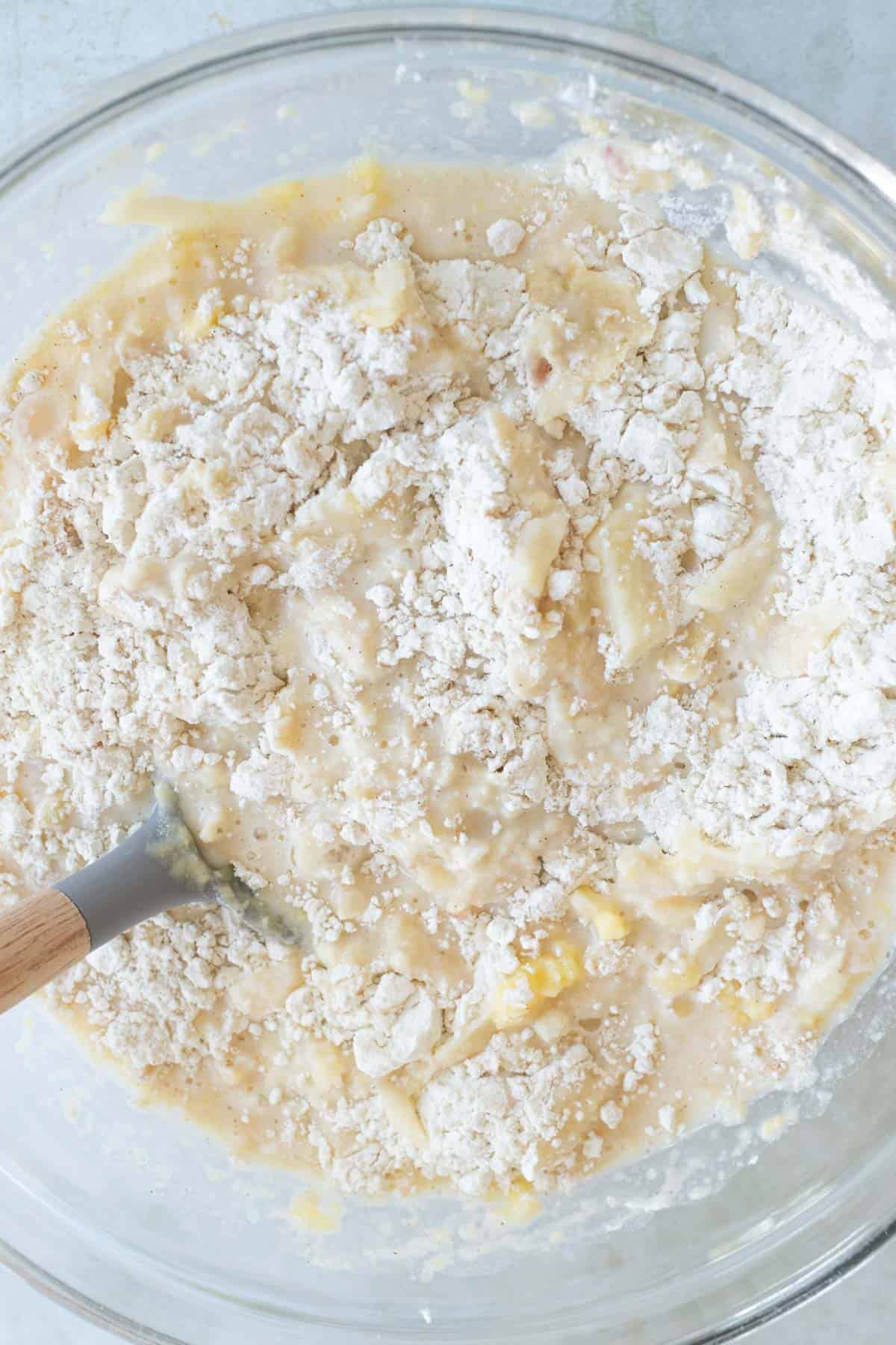 A mixing bowl with batter and partially mixed flour, featuring apple pancake batter and a wooden-handled spatula.