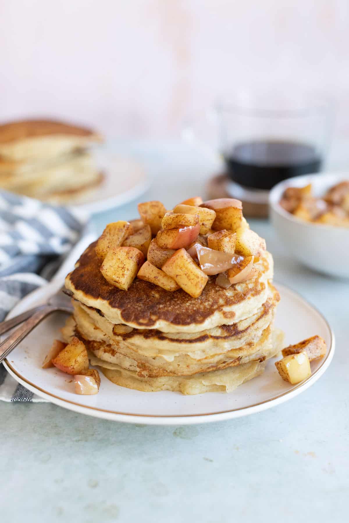 A stack of apple pancakes topped with diced apples and cinnamon on a white plate. A bowl of apple topping, a glass of coffee, and another plate of pancakes are in the background.
