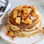 A stack of apple pancakes topped with cubed, spiced apples on a white plate with a fork and knife beside it.
