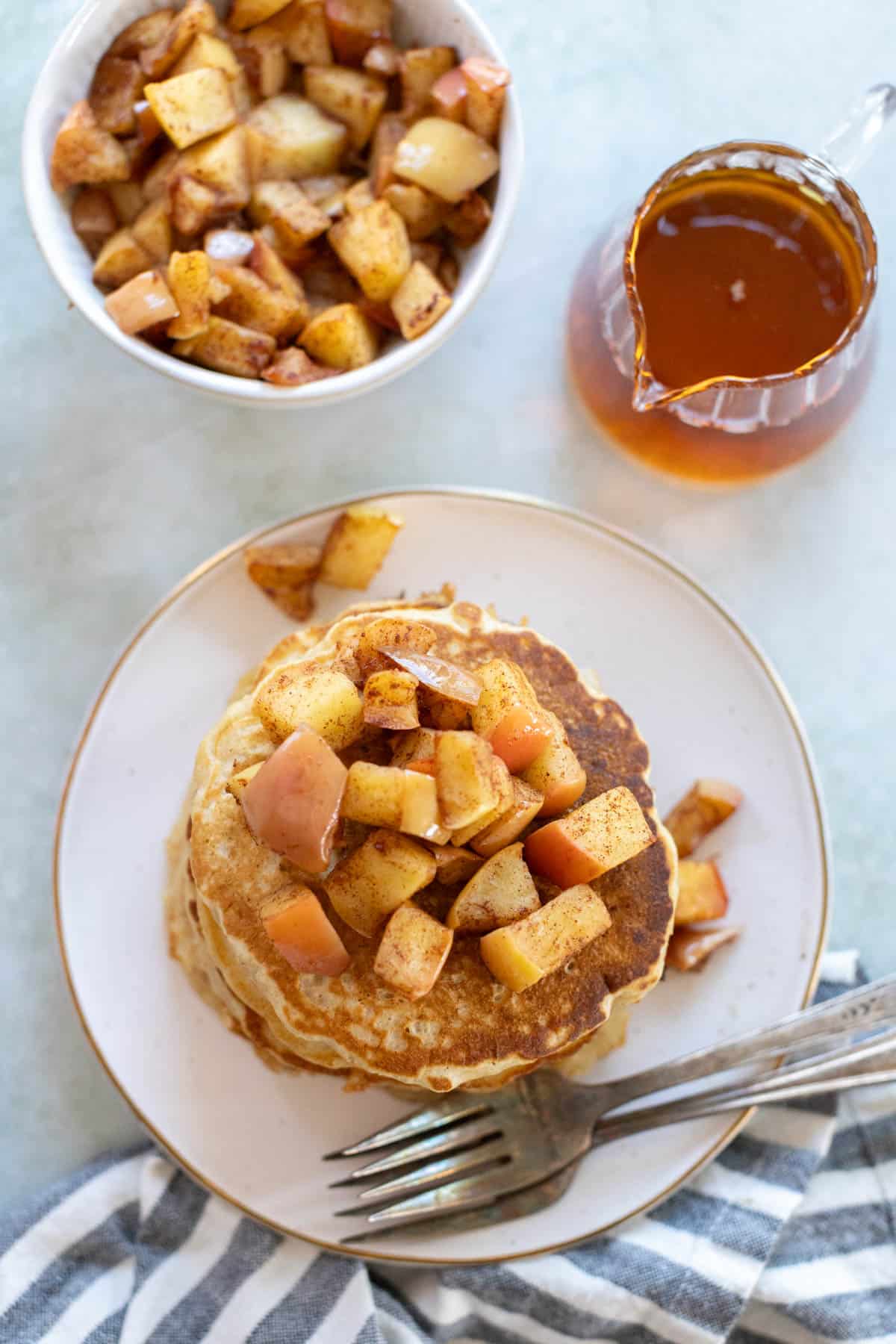 A plate of apple pancakes topped with diced apples next to a bowl of apples and a jug of syrup, with a striped cloth and utensils nearby.