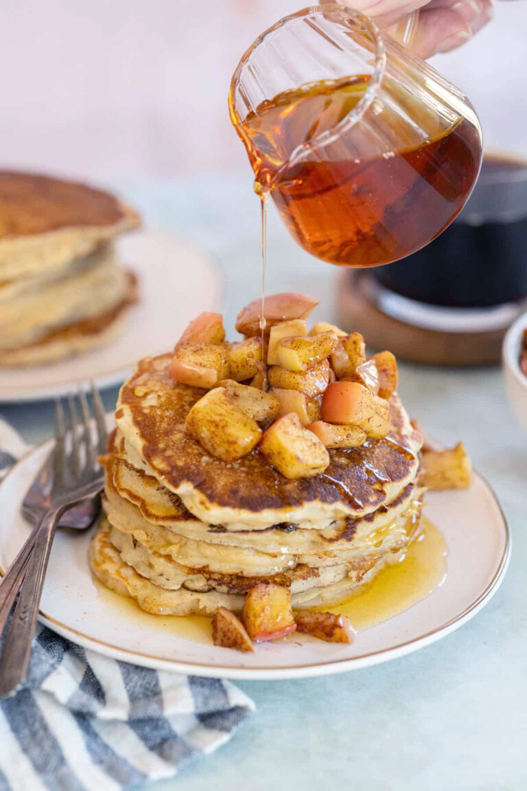A stack of apple pancakes topped with diced apples on a plate. Syrup is being poured from a glass container. A cup of coffee and a striped napkin are nearby.