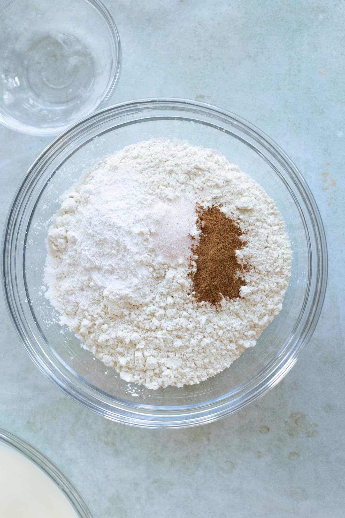 A glass bowl filled with flour, baking powder, baking soda, and salt on a textured light gray surface. Another small glass bowl and part of a larger bowl are partially visible nearby.