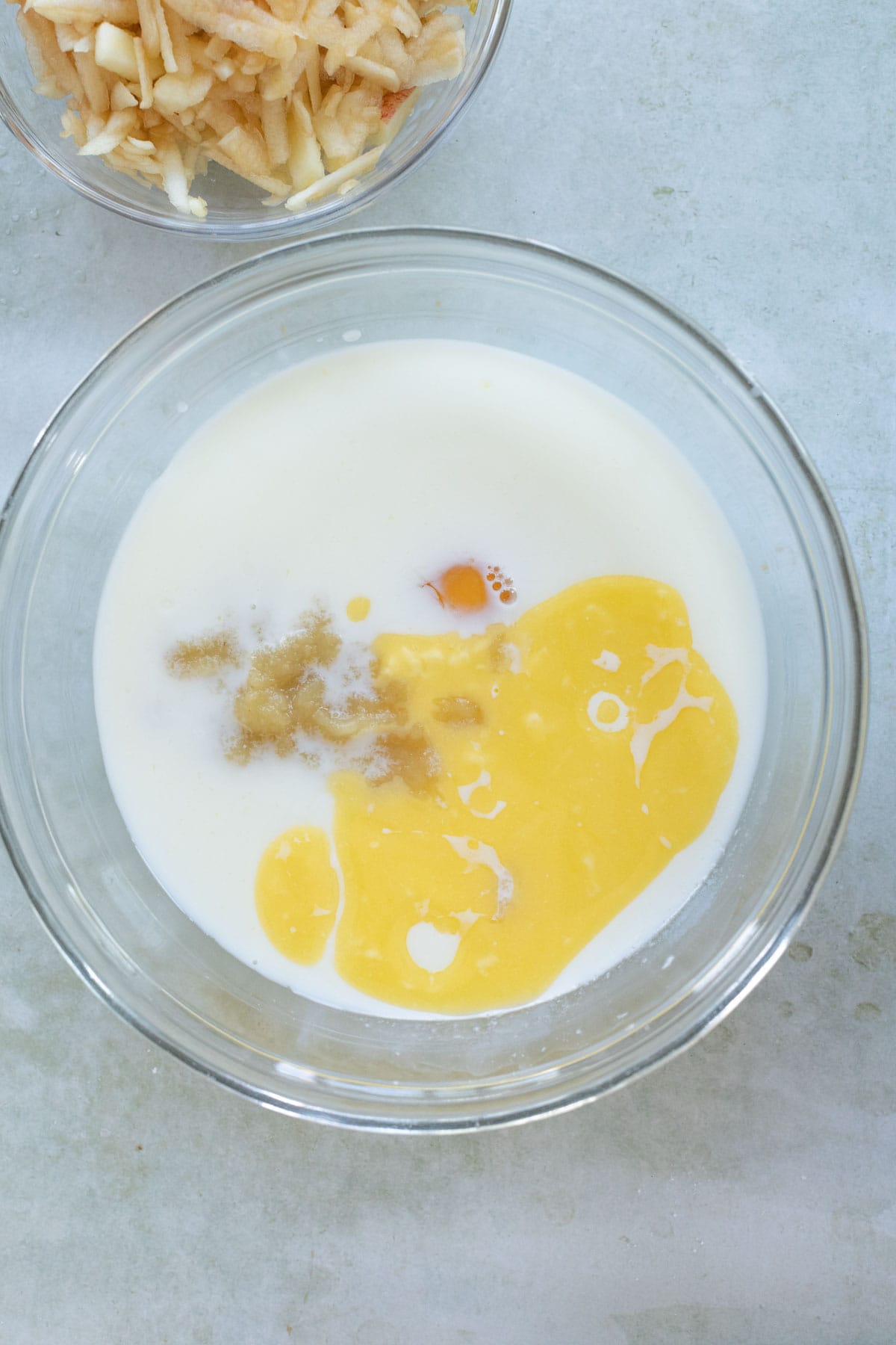 A glass bowl contains milk, an egg yolk, and applesauce. Another bowl with shredded apple is partially visible at the top.