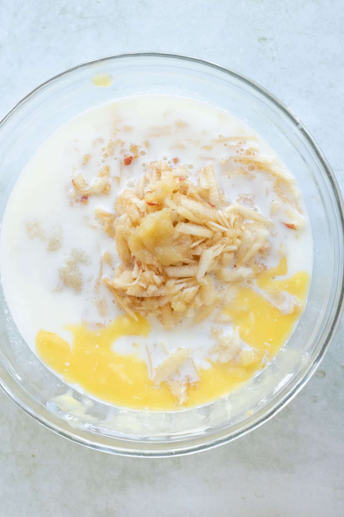 Glass bowl containing grated apples, milk, melted butter, and applesauce.