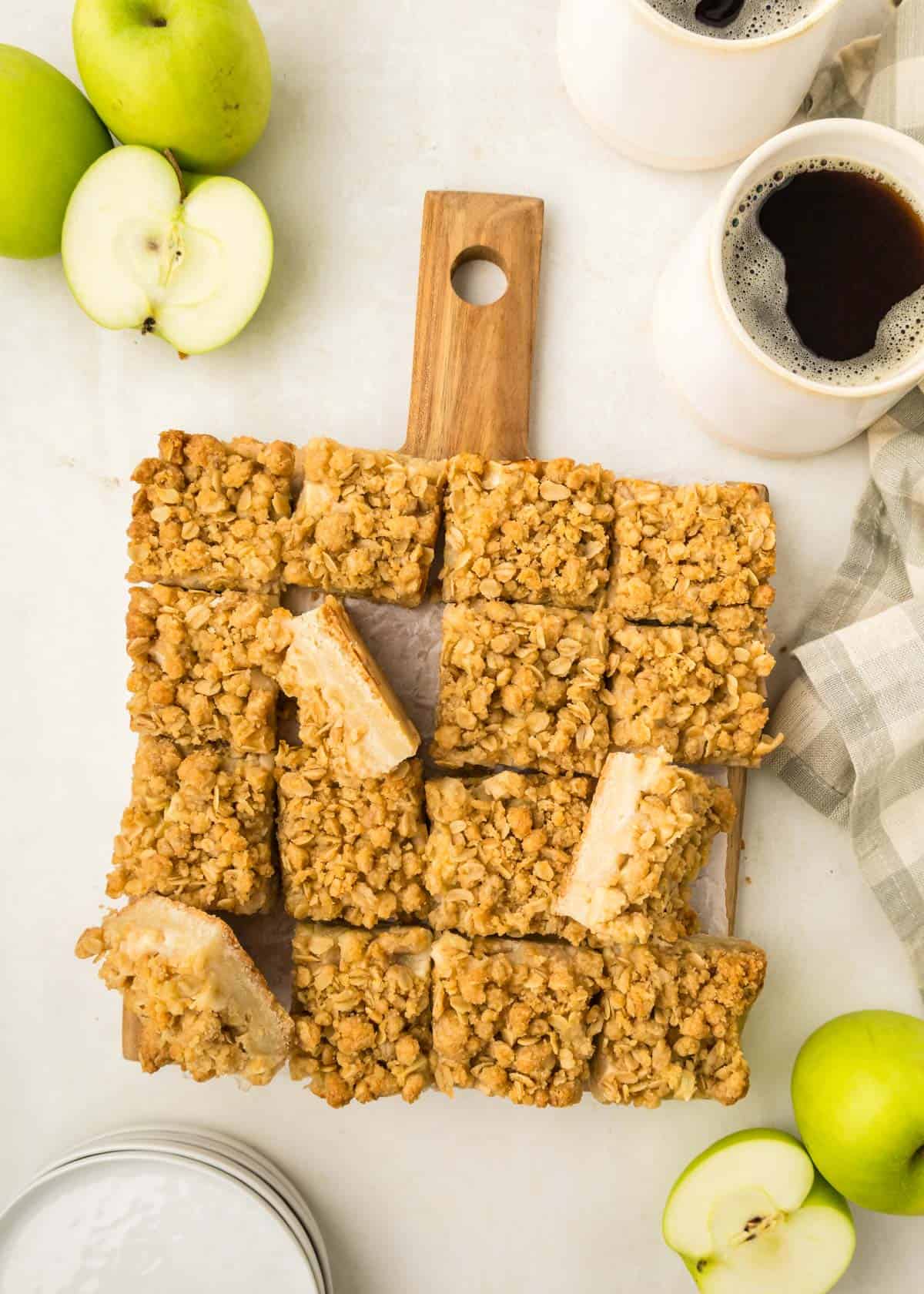 Sliced apple pie bars on a wooden board, surrounded by whole and cut green apples, two cups of coffee, and a striped cloth.