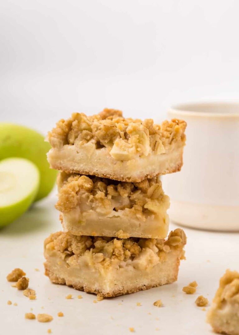 Three stacked apple pie bars with a crumbly topping, next to a whole green apple and a white mug.