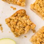 Four apple pie bars with a golden oat topping, arranged on a white surface, with a slice of green apple nearby.