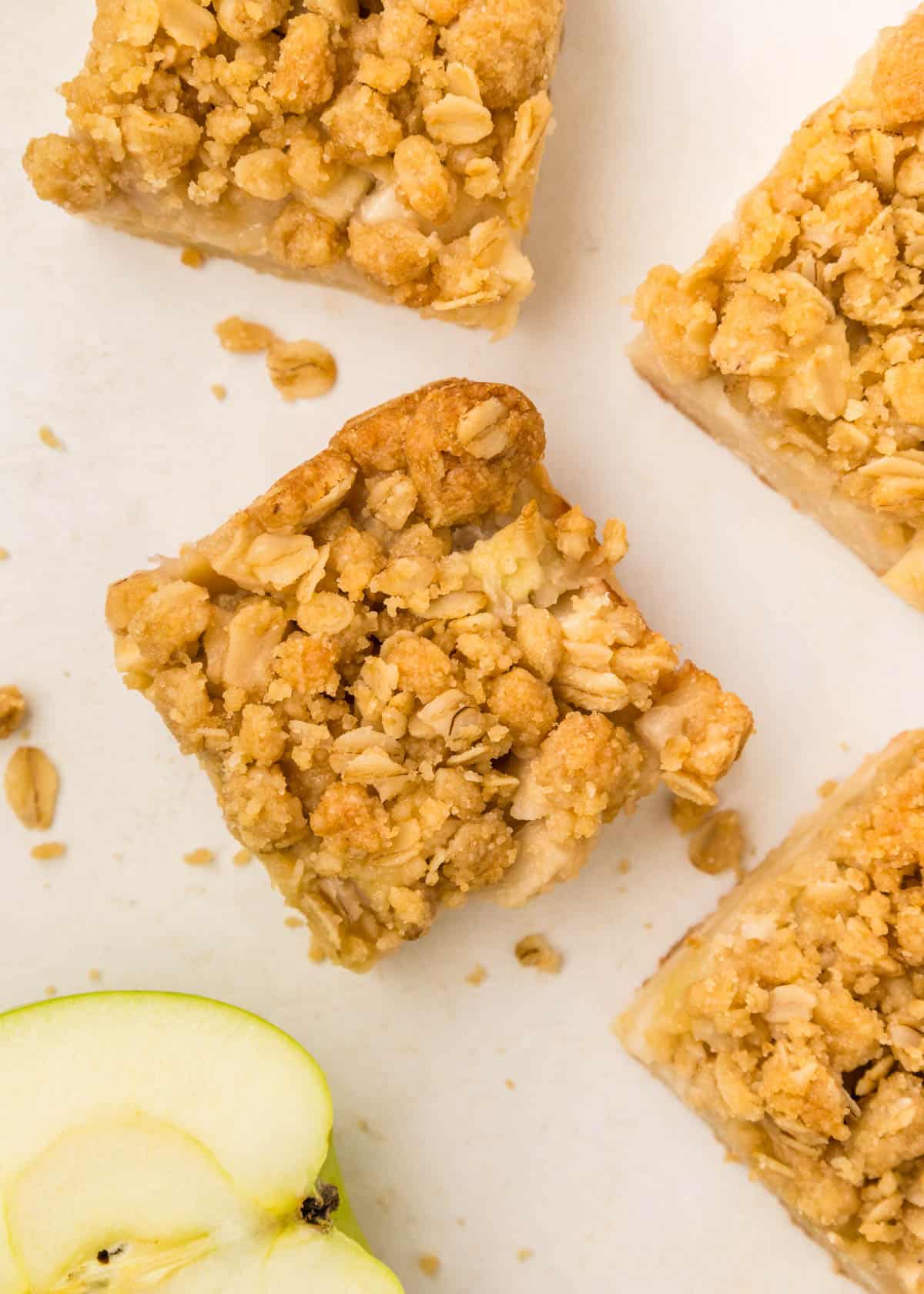Four apple pie bars with a golden oat topping, arranged on a white surface, with a slice of green apple nearby.