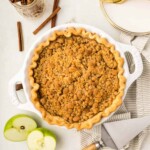A baking dish with apple crumble pie; a pie server, two apple halves, cinnamon sticks, a striped cloth, and gold utensils are nearby.