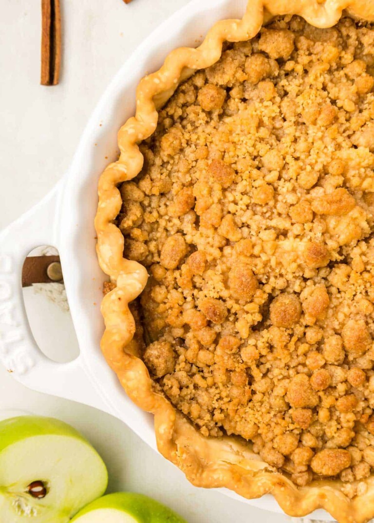 A baked apple crumble pie in a white dish, featuring a golden-brown crumb topping. Sliced green apples and a cinnamon stick are visible nearby.