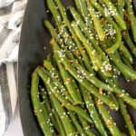 A plate of asian green beans garnished with sesame seeds and spices, placed next to a striped cloth.