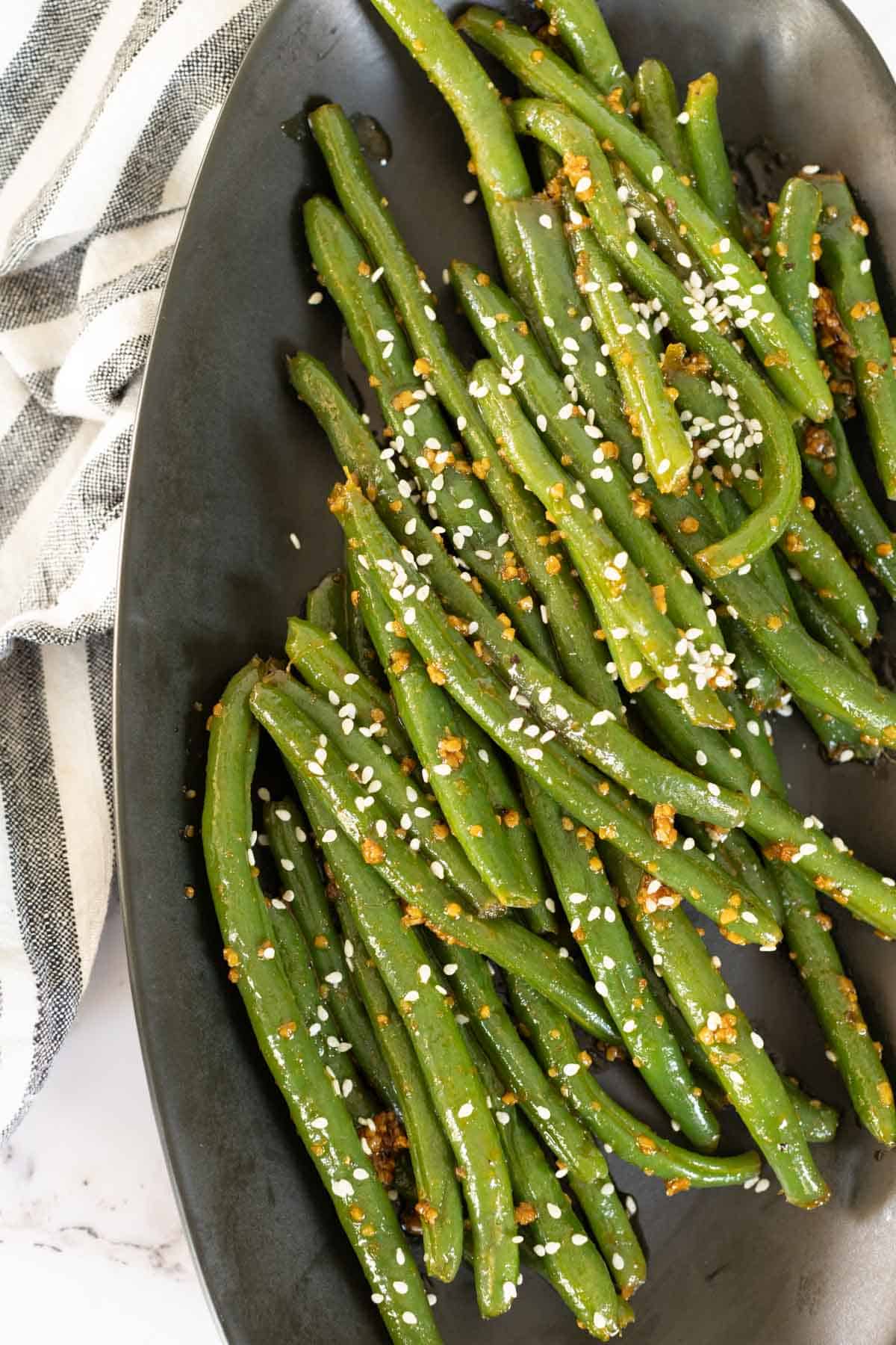 A plate of asian green beans garnished with sesame seeds and spices, placed next to a striped cloth.