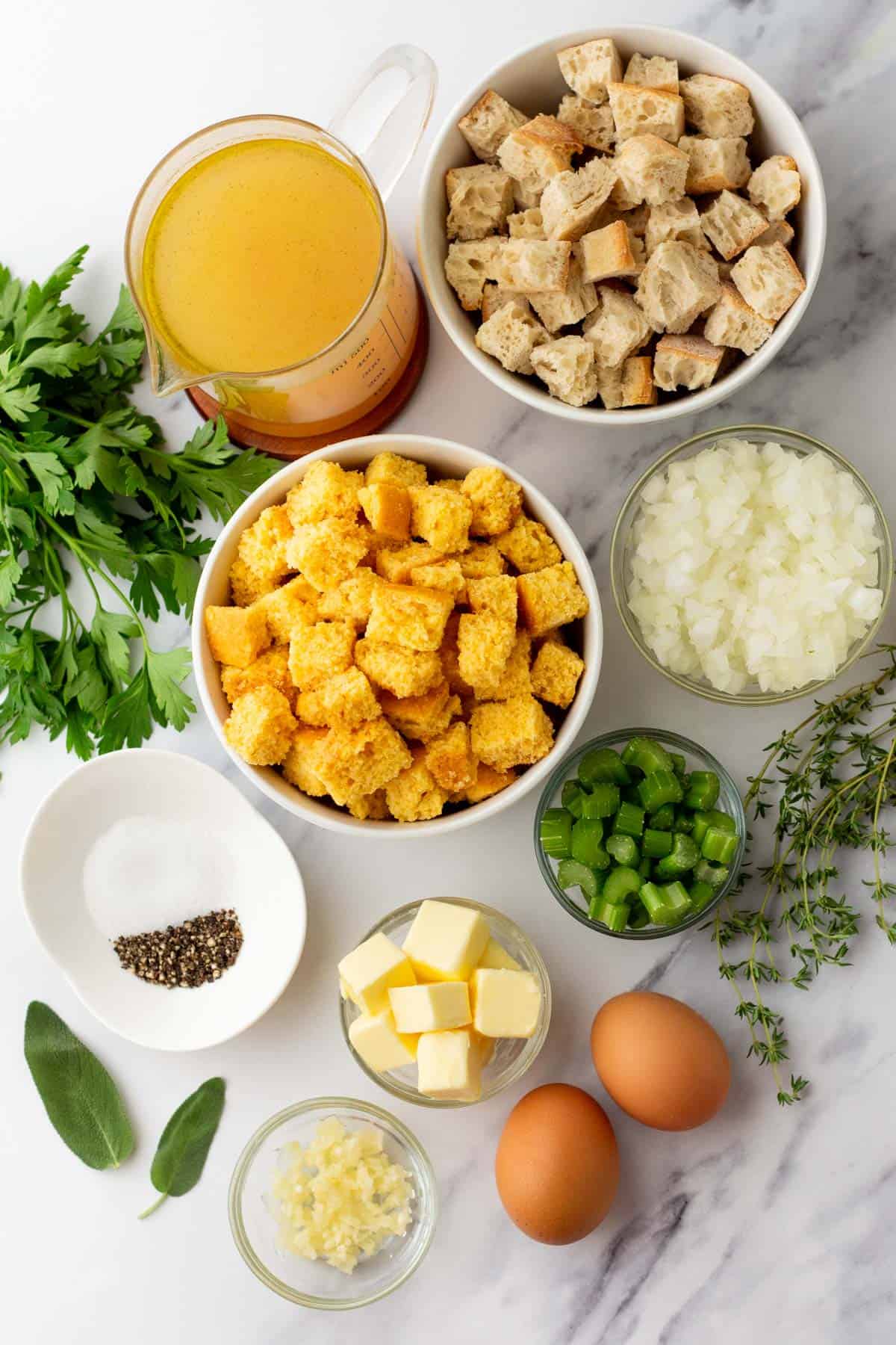 Ingredients on a marble surface: chopped parsley, broth, croutons, cornbread cubes, diced onions, diced celery, spices, butter, eggs, garlic, fresh herbs including thyme and sage.