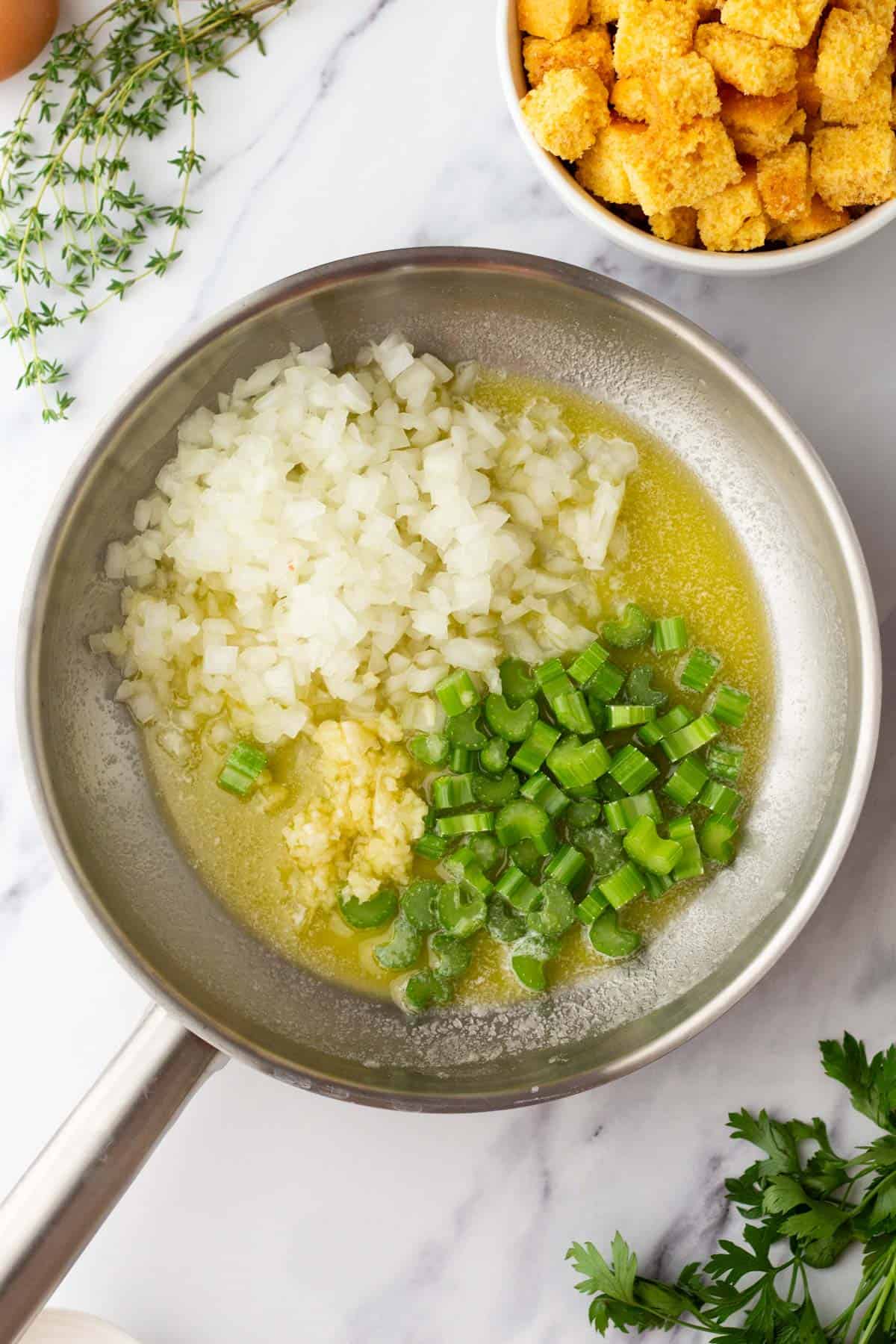A frying pan with chopped onions, celery, and minced garlic in butter. Fresh herbs are visible nearby, and a bowl of croutons is in the background.