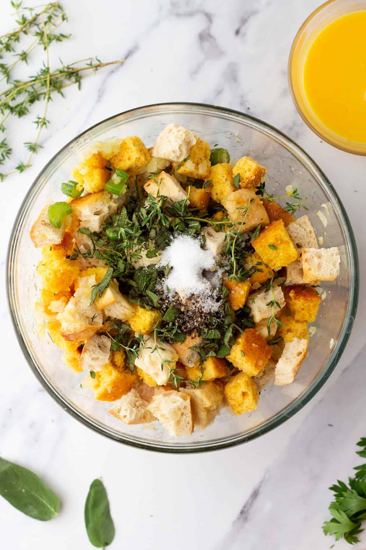 Glass bowl with cubed bread, herbs, and seasoning on a marble surface. Broth in a nearby glass bowl. Fresh herbs scattered around.