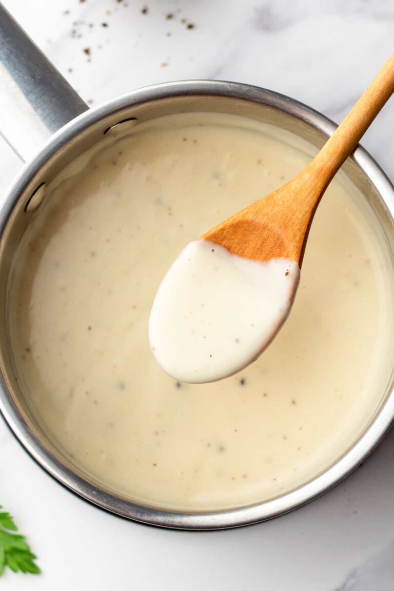 A wooden spoon holds a portion of cream of chicken above a saucepan placed on a marble surface, with some visible black pepper specks.