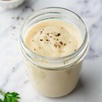 A jar of cream of chicken topped with cracked pepper, surrounded by parsley leaves and a spoon on a marble surface.