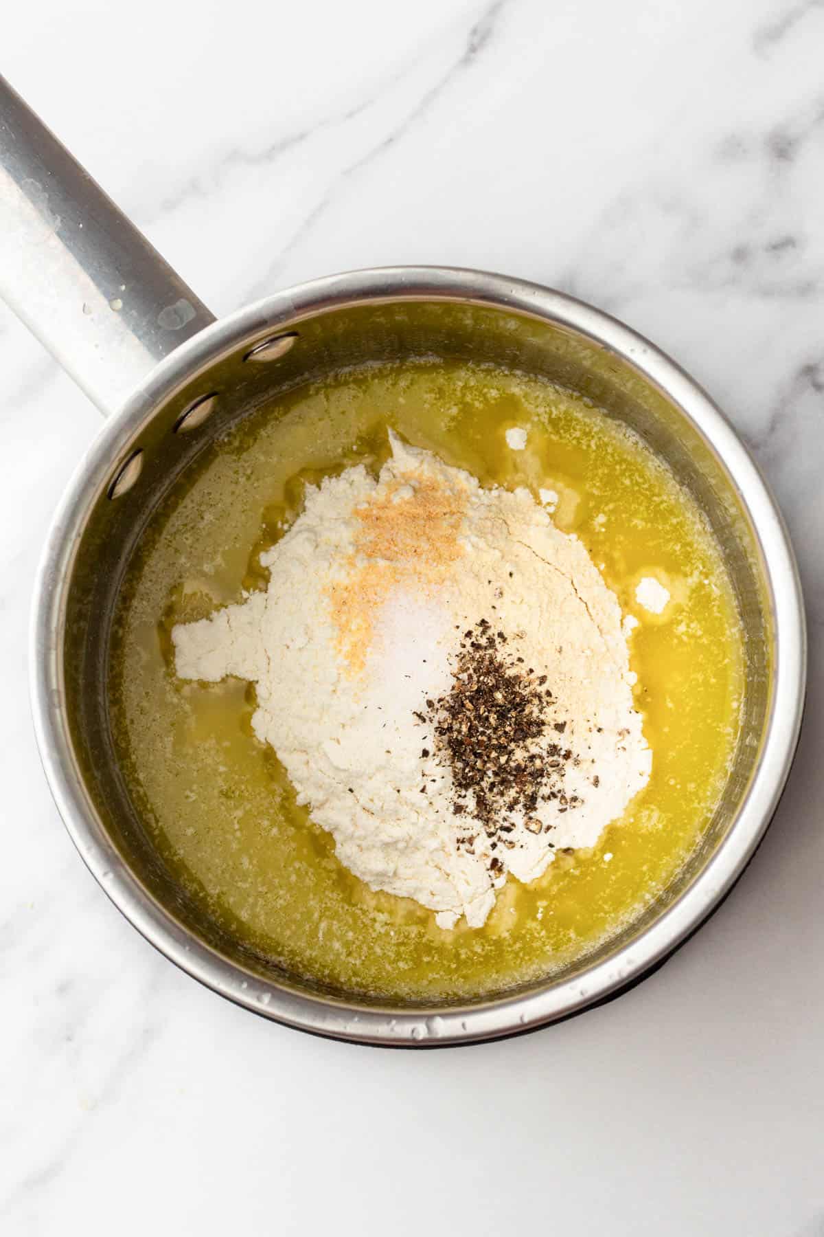 A saucepan with melted butter, flour, pepper, and salt on a white marble surface.