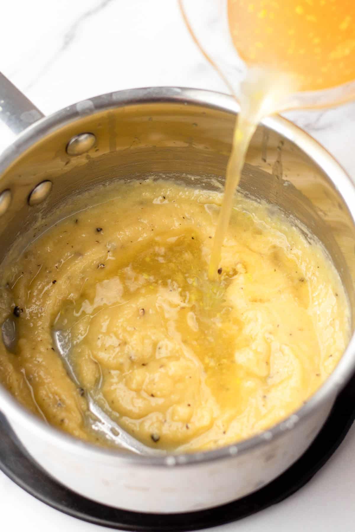 Pouring broth into a saucepan with a creamy base on a stove.