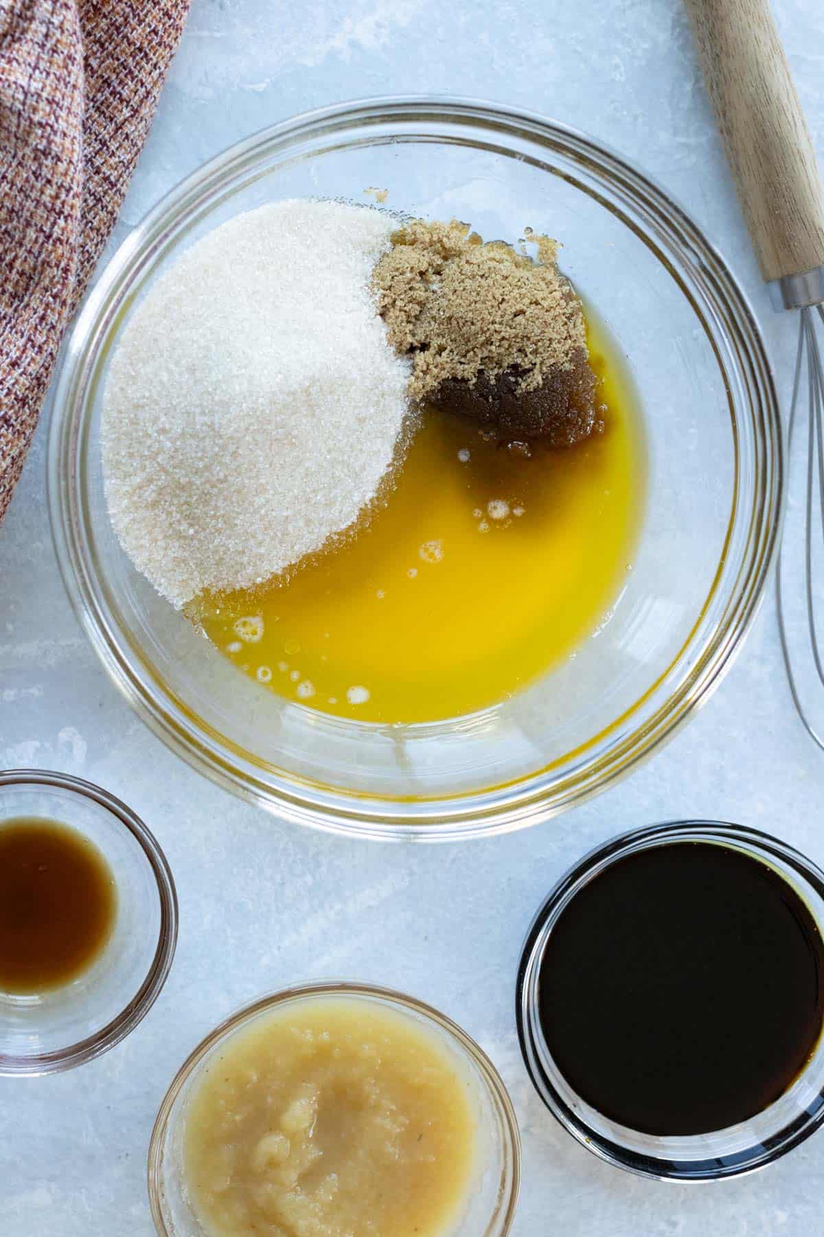 A bowl with melted butter, white sugar, and brown sugar. Nearby are small bowls containing vanilla extract, applesauce, and molasses on a light countertop. A whisk is partially visible.