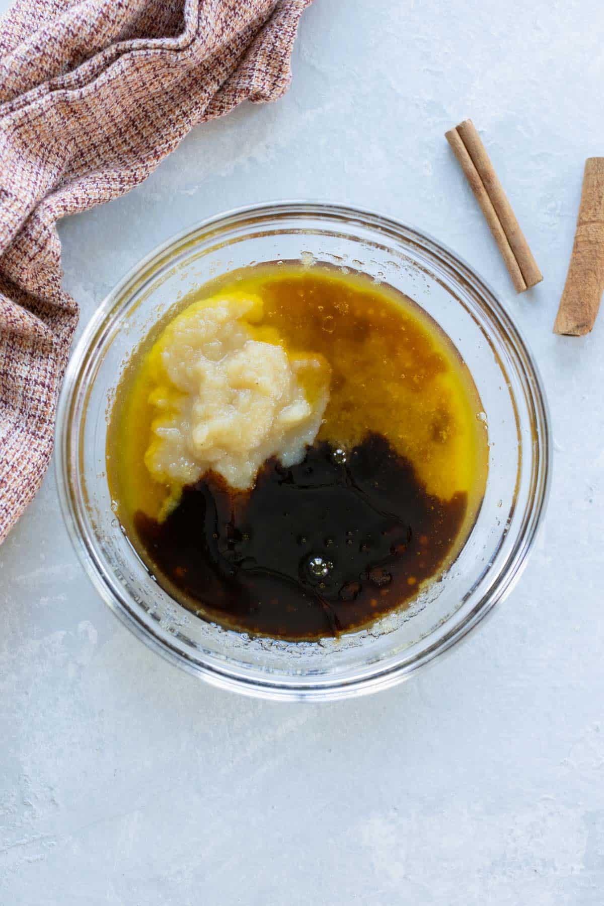 A glass bowl contains melted butter, applesauce, and vanilla on a light surface. A cloth and cinnamon sticks are nearby.