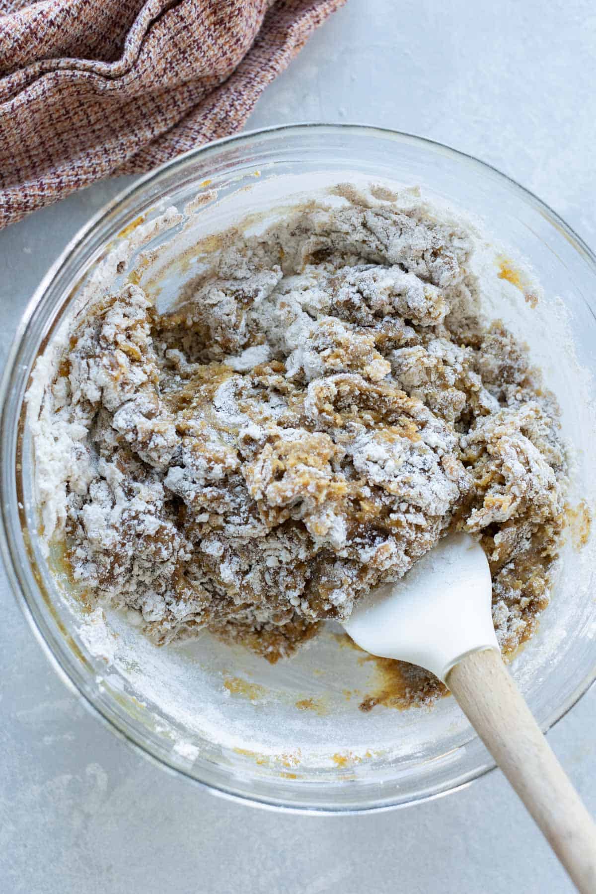 A mixing bowl contains raw ginger cookie dough with a white spatula, partially mixed with flour visible on top. A brown cloth is in the background on a gray surface.