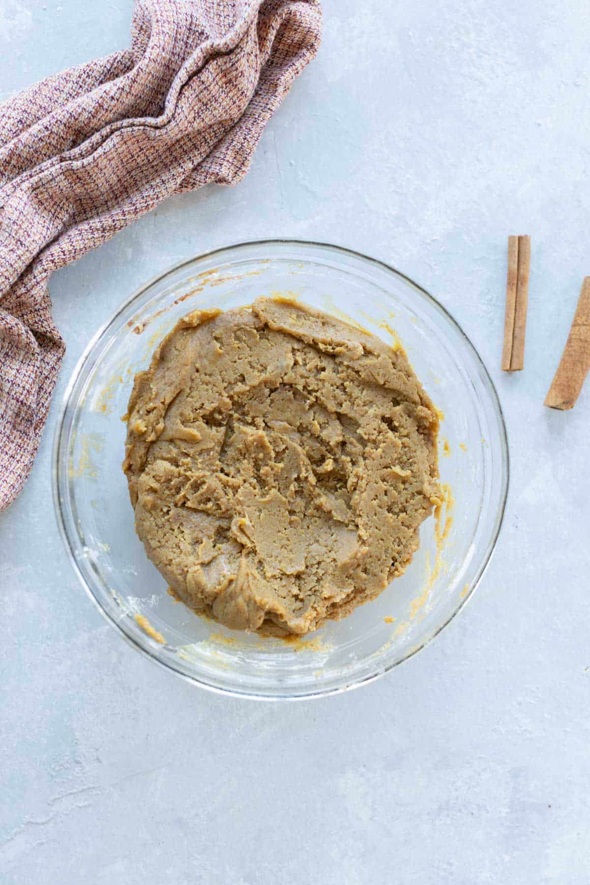 A glass bowl containing ginger cookie dough on a light surface, with a cloth and two cinnamon sticks nearby.