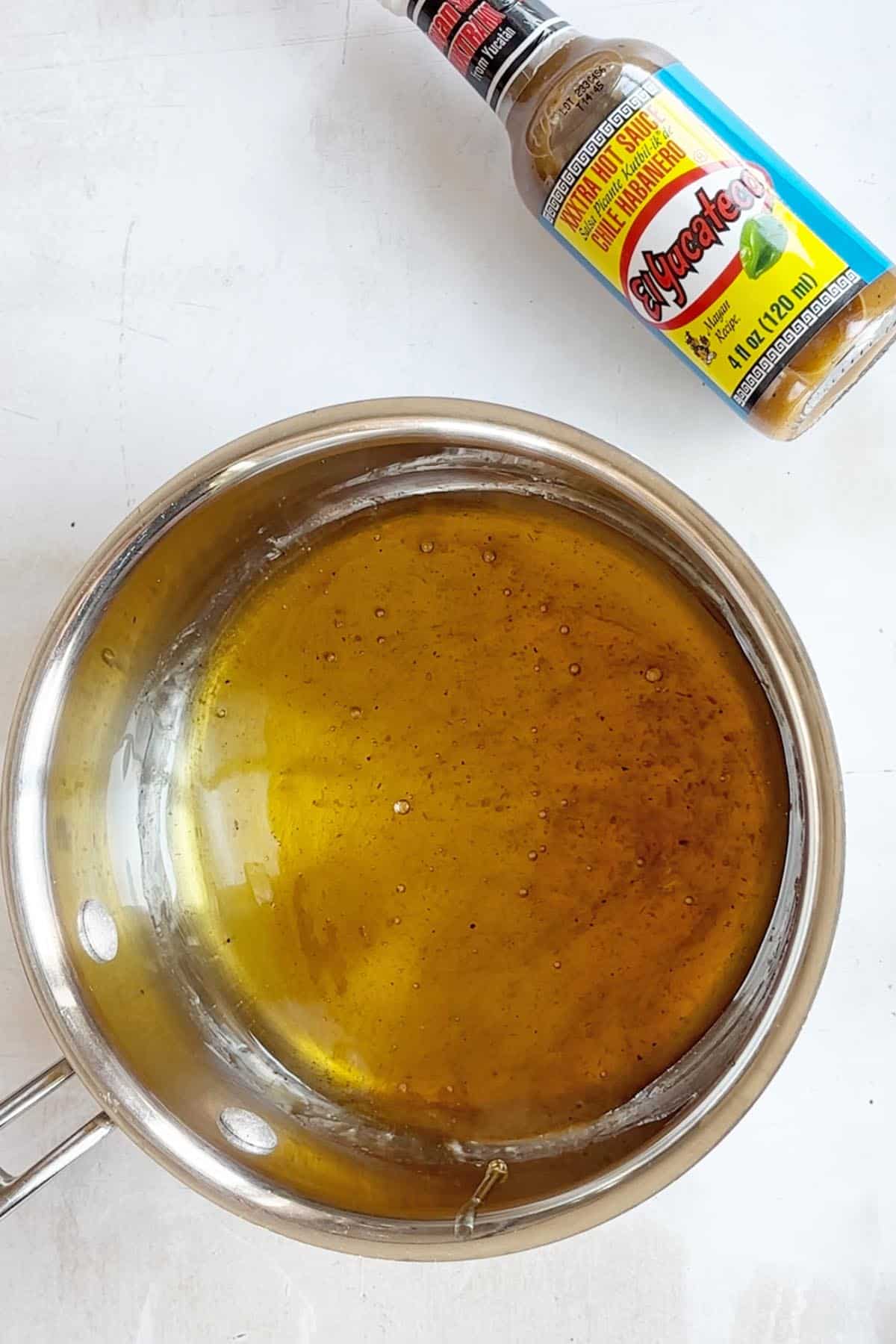 A small pot filled with a yellow, seasoned liquid sits on a white surface. A bottle labeled "Aygechew" is positioned at the top, hinting at the intriguing blend of flavors like habanero honey that lie within.