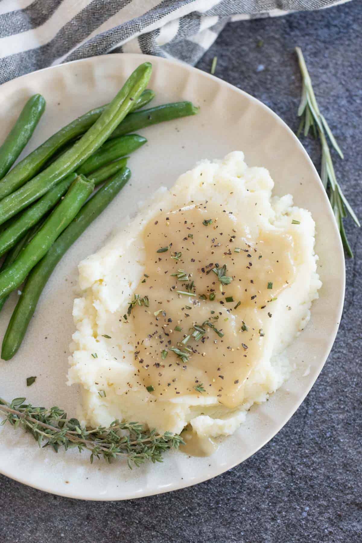 Plate of mashed potatoes with gravy, garnished with herbs, served alongside green beans on a light-colored dish.