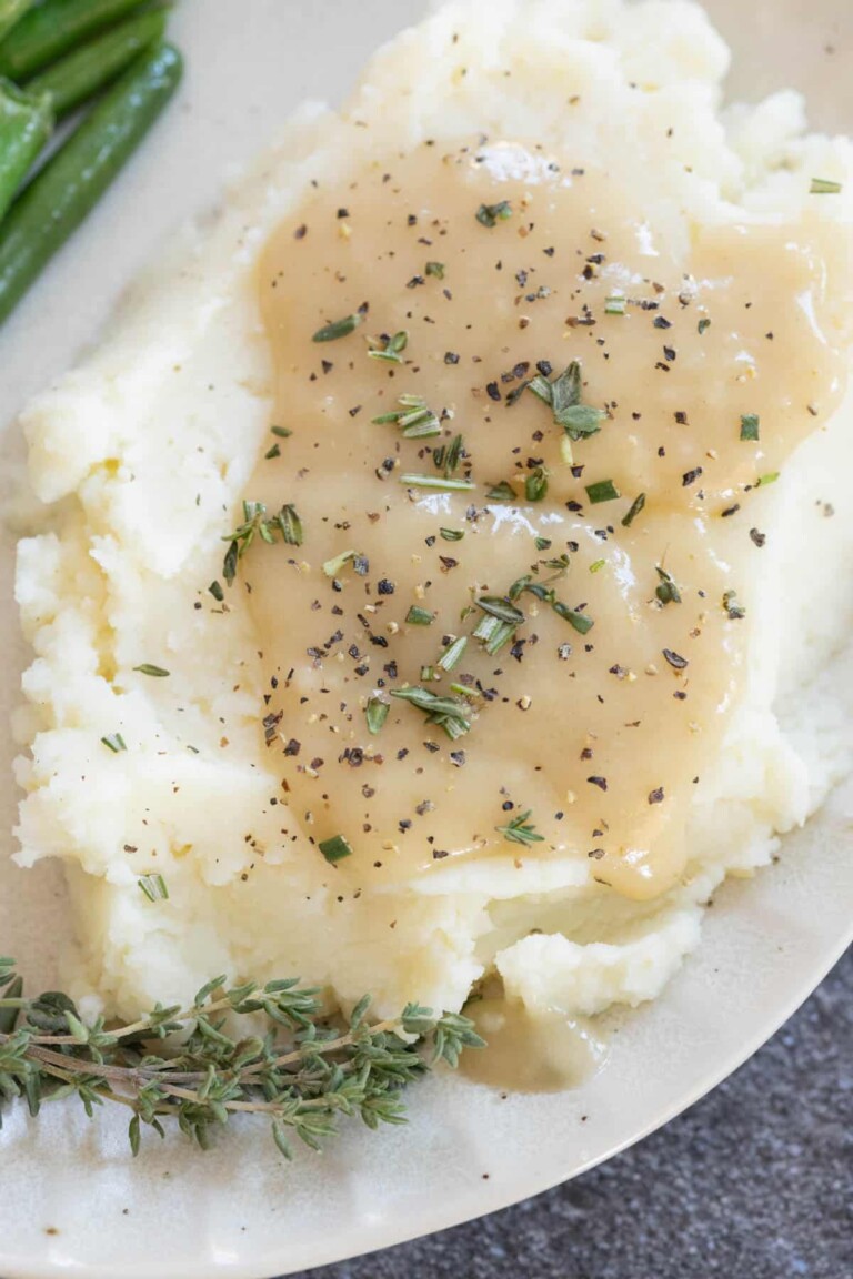 Mashed potatoes topped with gravy and sprinkled with herbs, served with a side of green beans and fresh thyme on a plate.