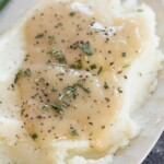 Plate of mashed potatoes topped with gravy and sprinkled with herbs and black pepper.