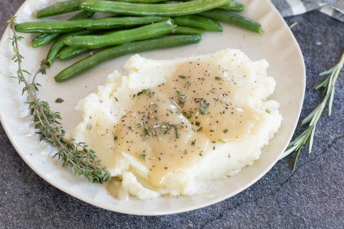 Plate with mashed potatoes topped with gravy, garnished with herbs beside green beans and a sprig of rosemary on a textured surface.