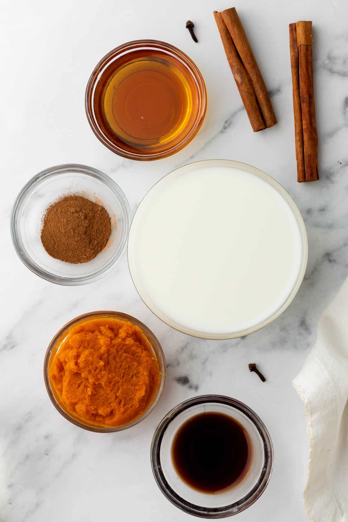 A collection of ingredients including a bowl of milk, a bowl of pumpkin puree, a small bowl of cinnamon, a small bowl of vanilla extract, a bowl of honey, two cinnamon sticks, and two cloves on a white surface.