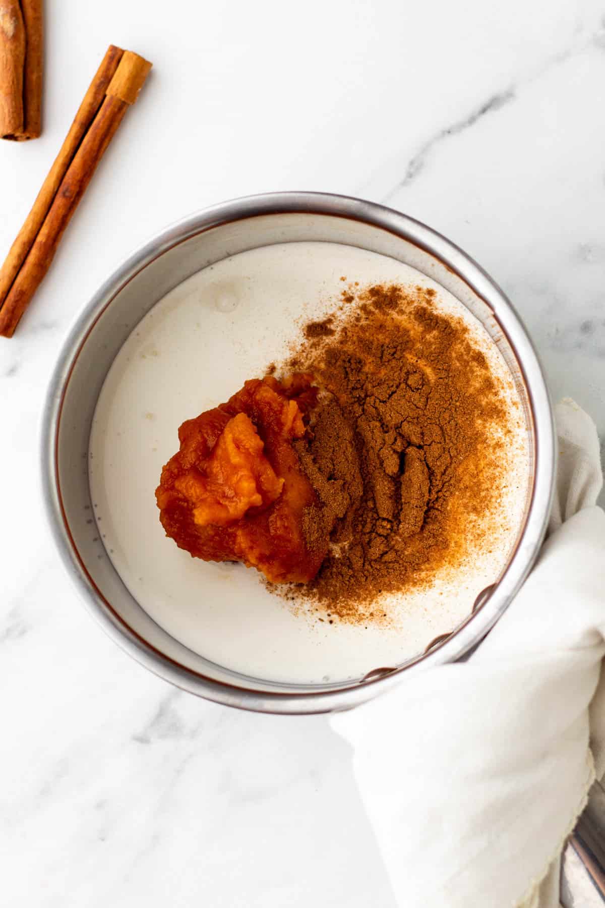 Overhead view of a bowl with milk, pumpkin pur&eacute;e, and cinnamon on a marble surface. Two cinnamon sticks are placed nearby.