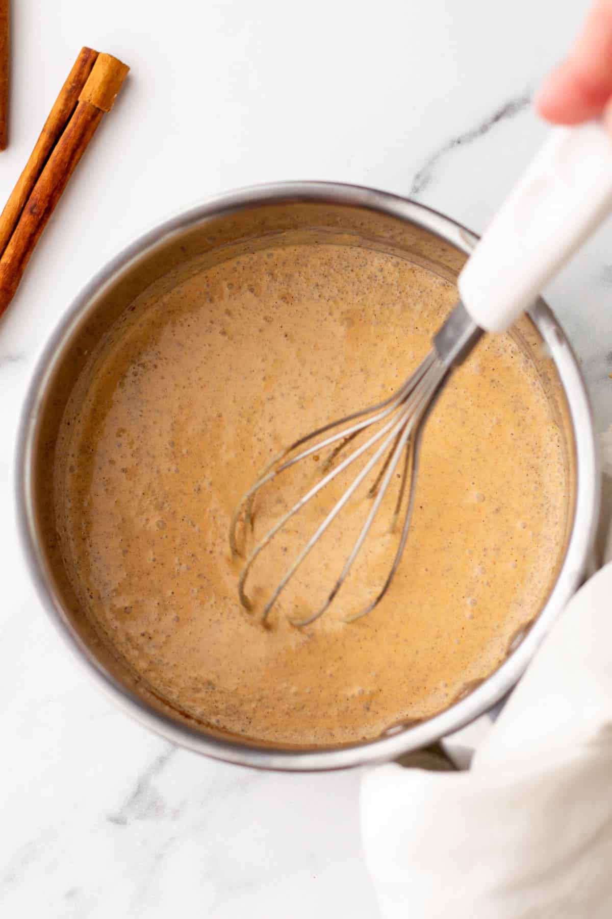 A whisk stirring a light brown mixture in a round metal bowl, with cinnamon sticks beside it on a marble countertop.