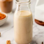 A glass bottle filled with a creamy beige liquid is placed on a marble surface. Nearby are cinnamon sticks, cloves, and a bowl of brown paste in the blurred background.