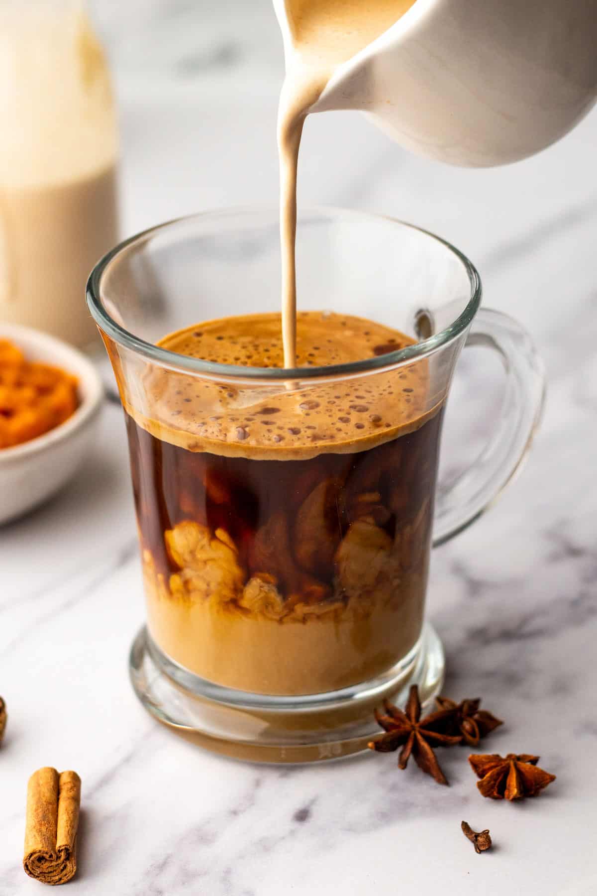 Cream is being poured into a glass mug filled with coffee, surrounded by cinnamon sticks and star anise on a marble surface.