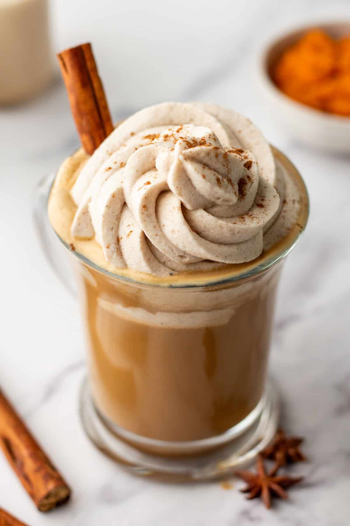 A glass mug of pumpkin latte topped with whipped cream and a sprinkle of cinnamon, garnished with a cinnamon stick. In the background, a bowl of pumpkin puree and another cinnamon stick are visible.
