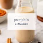 A sleek glass bottle of pumpkin creamer rests on a marble surface, accompanied by aromatic cinnamon sticks and a softly blurred bowl in the background.