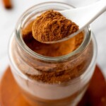 A spoonful of pumpkin pie spice is held above an open glass jar filled with more cinnamon, placed on a wooden coaster.