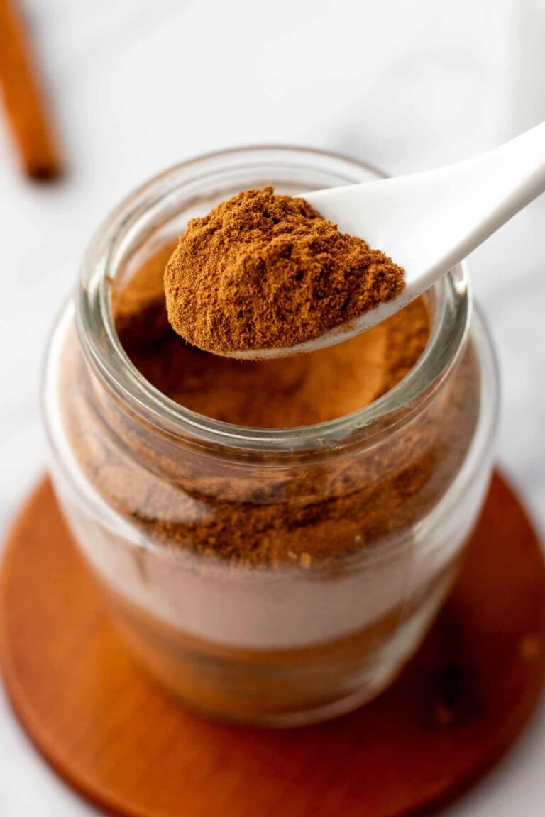 A spoonful of pumpkin pie spice is held above an open glass jar filled with more cinnamon, placed on a wooden coaster.